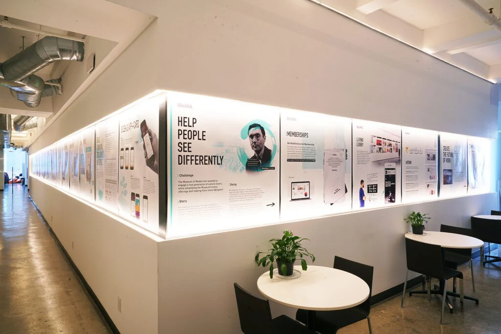 Co-Collective: Illuminated project display wall beside small round tables and black chairs